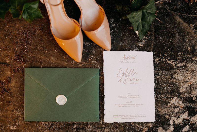 Flatlay%20met%20oranje%20hakken%2C%20een%20donkergroene%20envelop%20met%20witte%20waxzegel%20en%20een%20trouwuitnodiging%20op%20wit%20papier